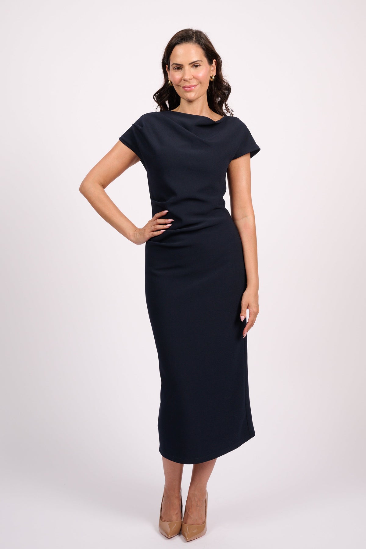A woman modeling a short-sleeve, knee-length navy dress with a draped neckline, standing against a light gray studio background.