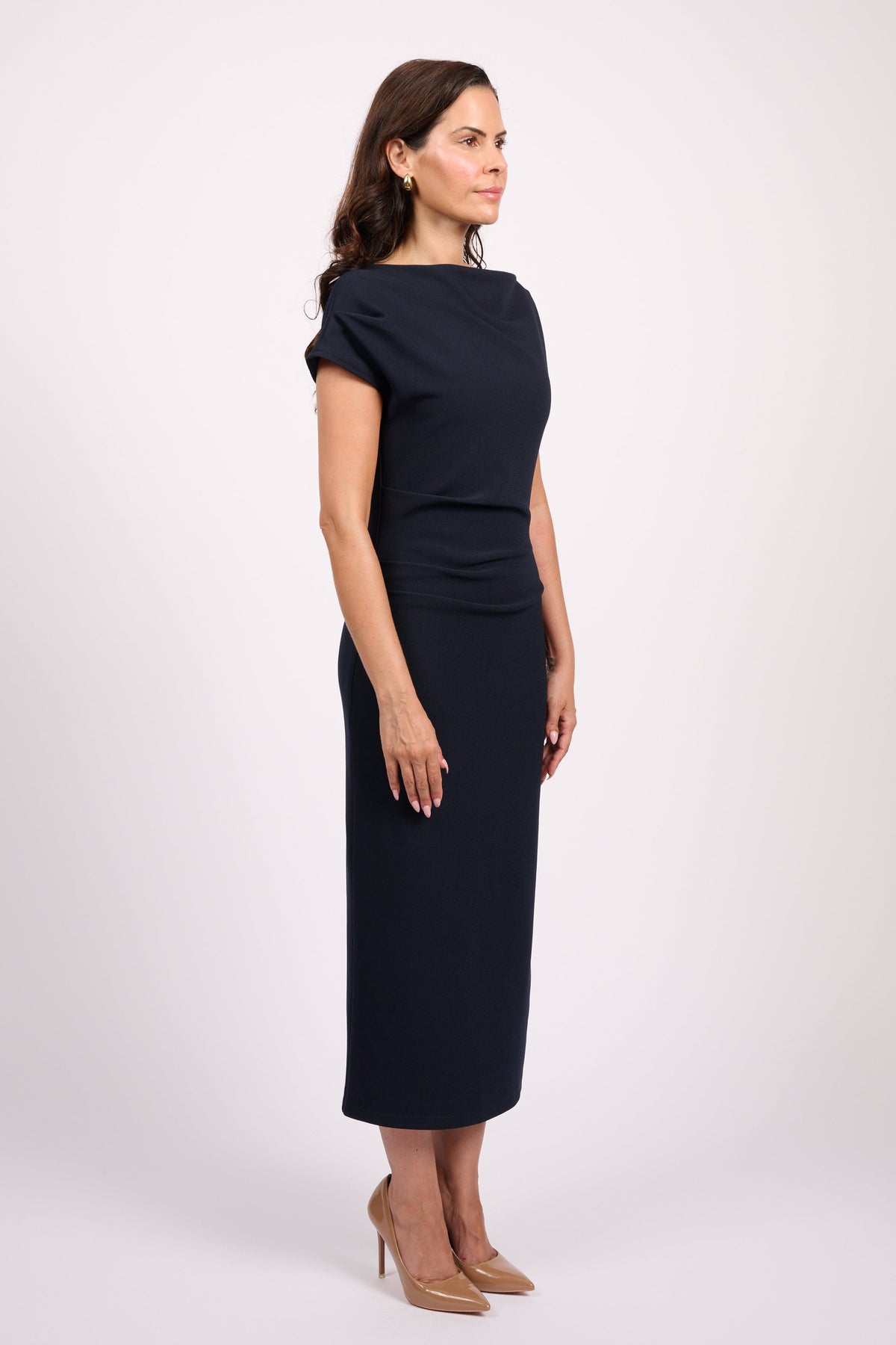 A woman modeling a dark navy midi dress with short sleeves and a boat neckline, standing in profile against a light gray studio background.