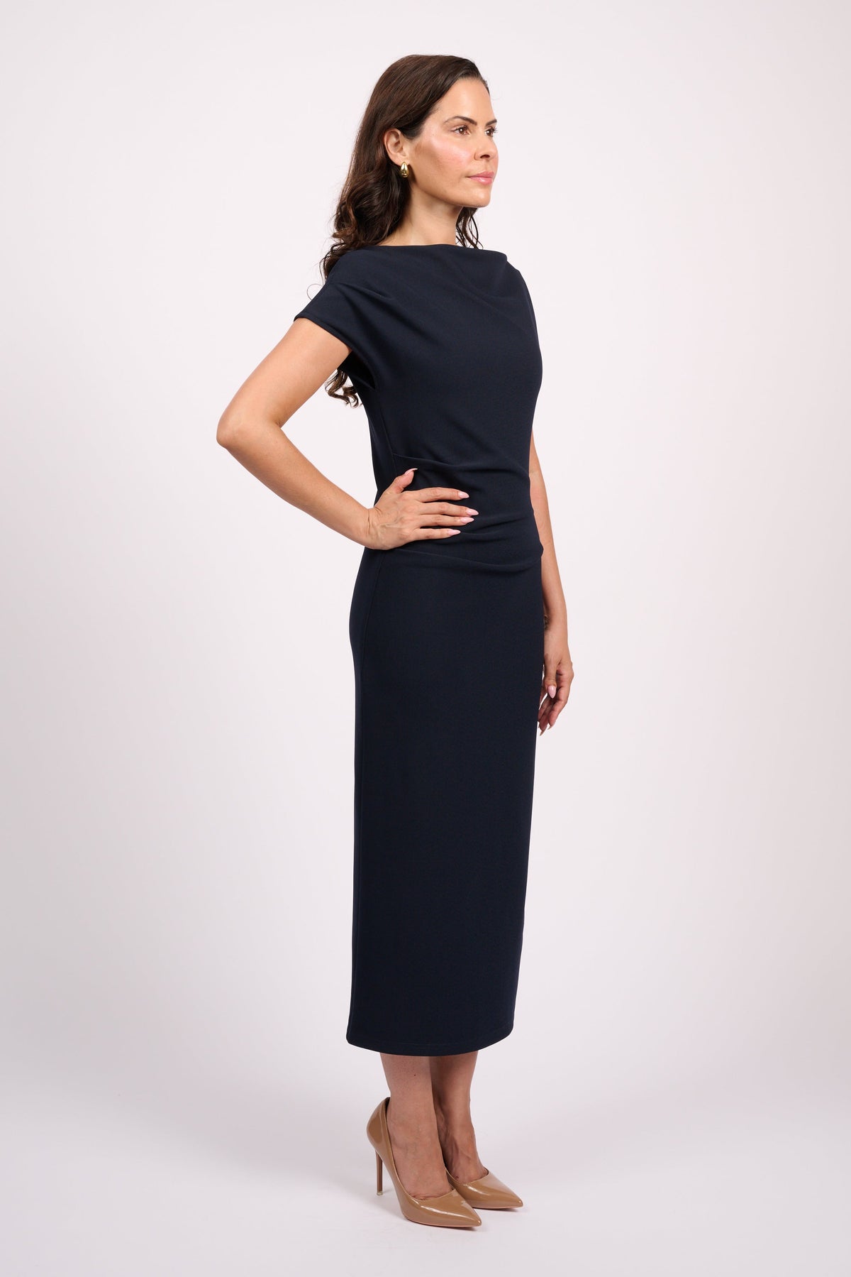A woman modeling a dark navy midi dress with short sleeves and a boat neckline, standing in profile against a light gray studio background.