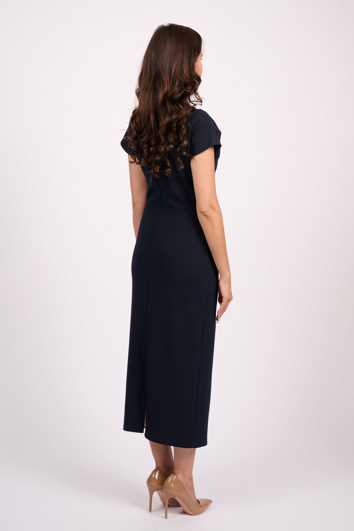 A woman is shown from the back wearing a dark navy midi dress with short cap sleeves and nude high heels, standing against a plain light gray studio background.