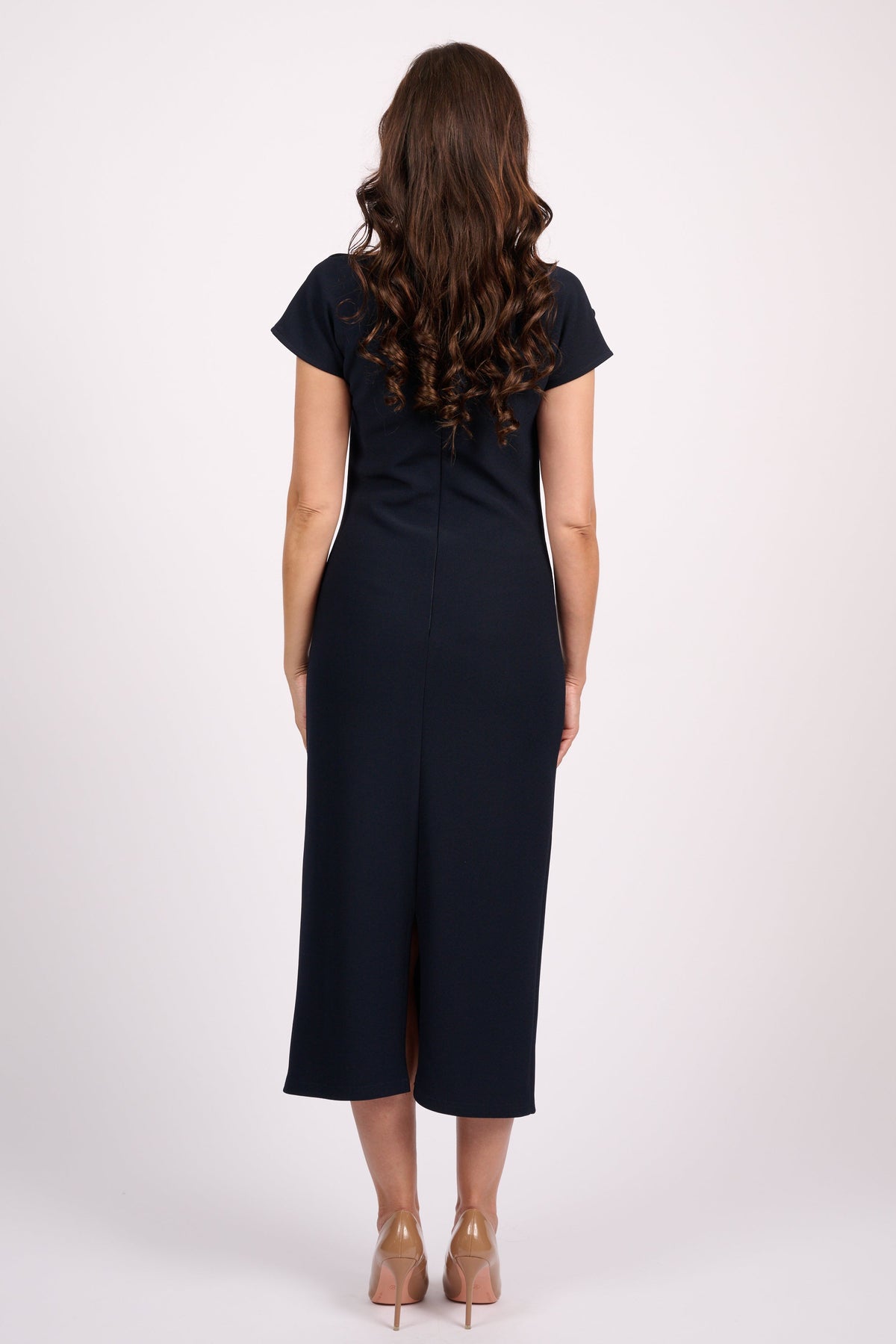A woman viewed from the back wearing a dark navy midi dress with short sleeves and nude high heels, standing against a plain light gray studio background.