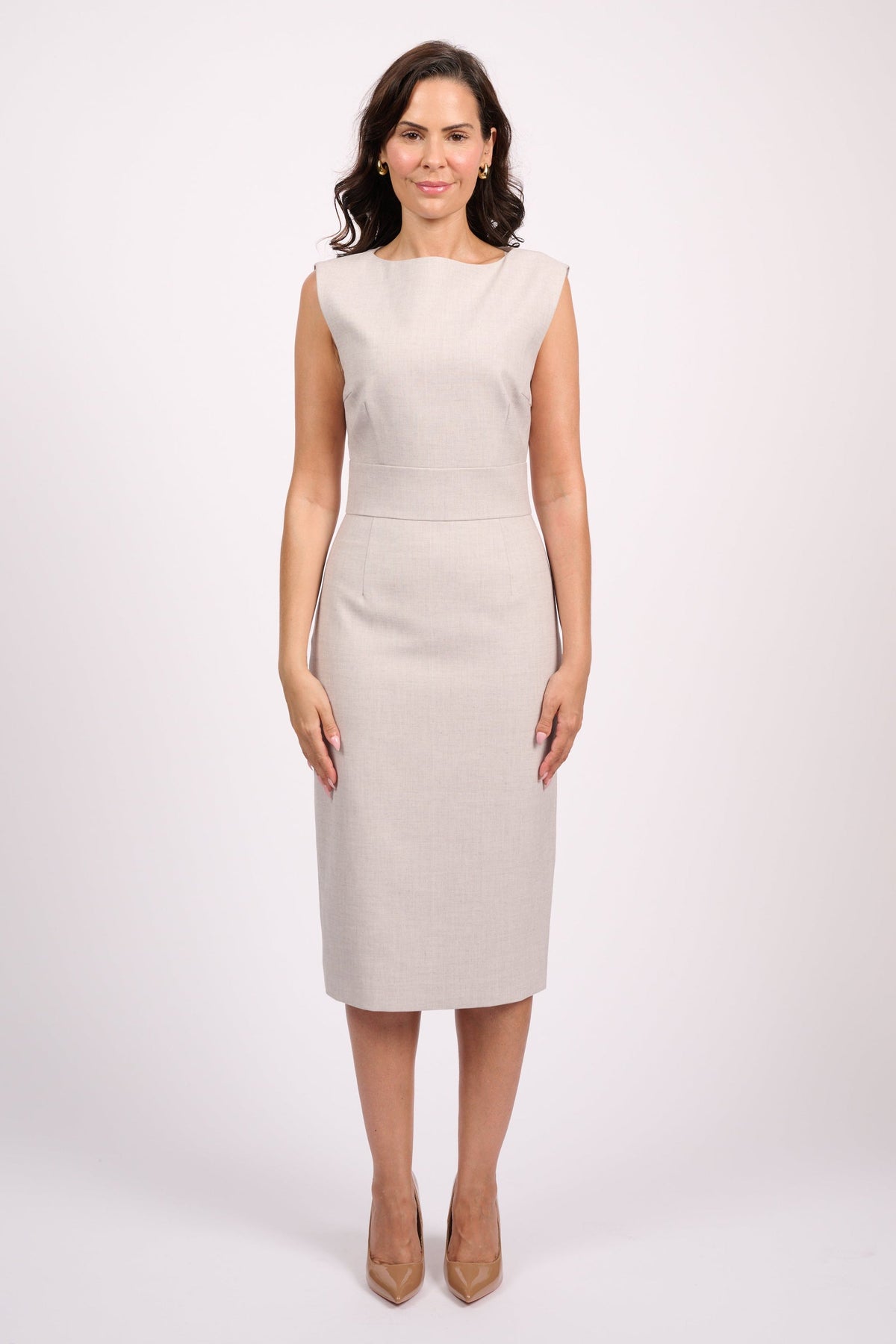 A woman modeling a sleeveless, knee-length beige dress with structured shoulders and a straight silhouette, paired with beige heels, against a plain light gray studio background.