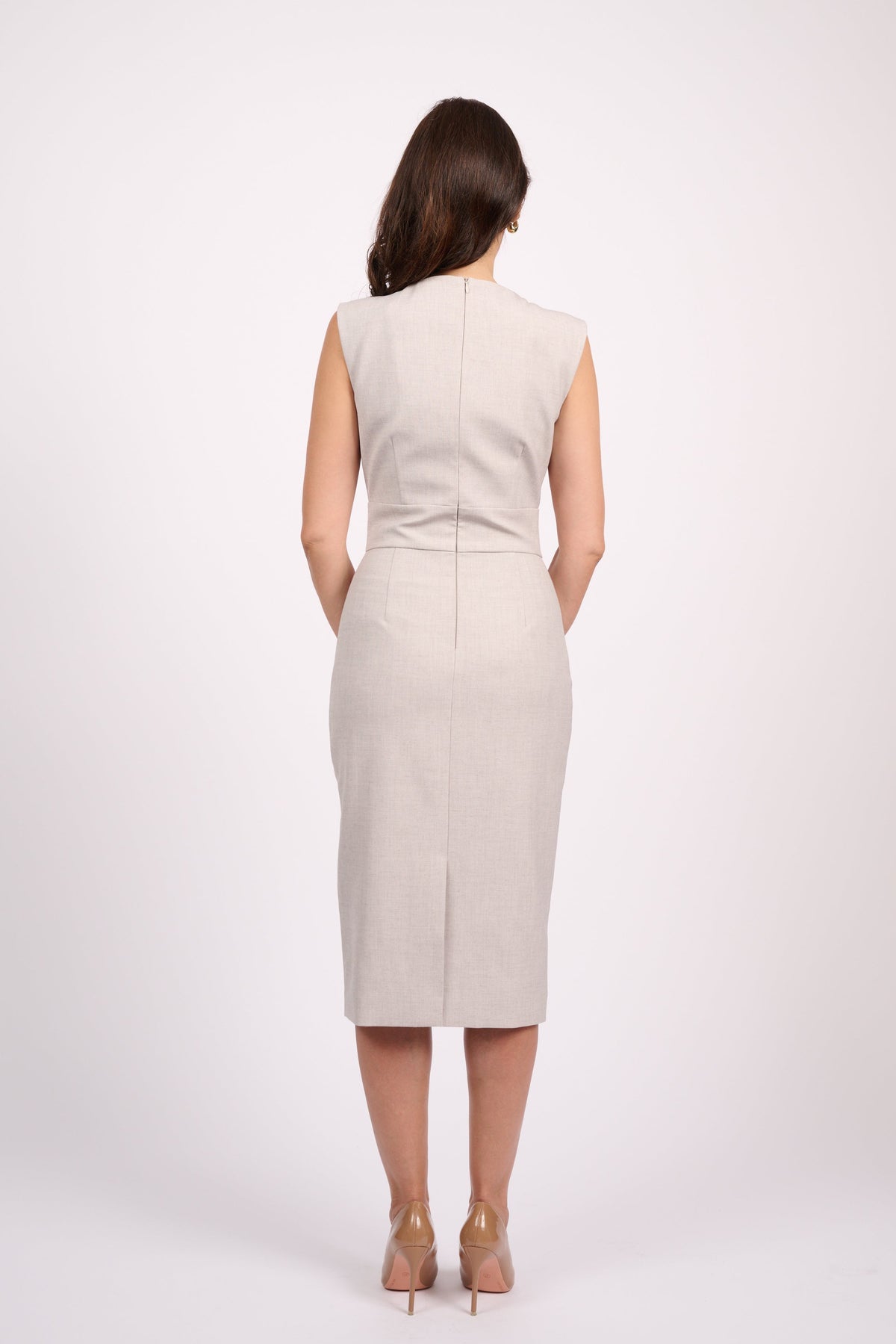A woman viewed from the back wearing a sleeveless, knee-length beige dress with a straight neckline and a front center seam, paired with nude high heels, against a plain light gray studio background.